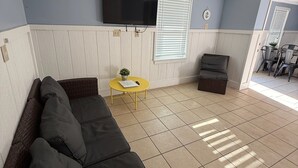 Uma sala de estar com um sofá de cor escura, uma mesinha amarela e uma televisão de tela plana fixada na parede.