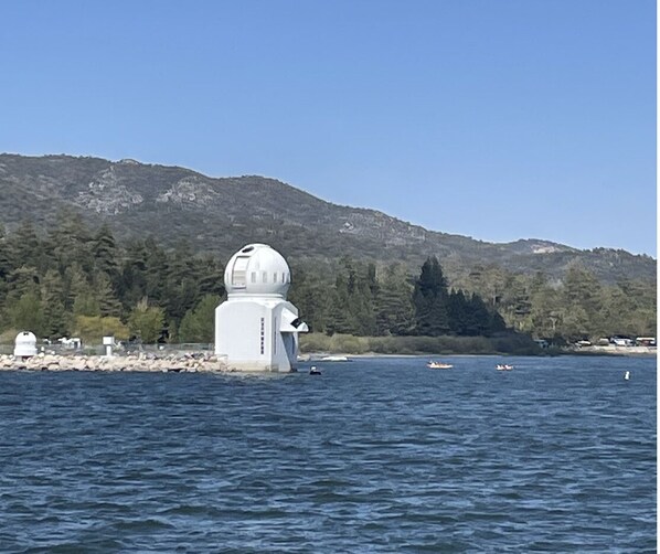 Un edificio blanco con una cúpula a la orilla de un lago, rodeado de aguas tranquilas y una ladera cubierta de bosque.