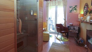 Una habitación con interior de madera, mampara de vidrio en la ducha, una mecedora, una mesita con objetos decorativos y una ventana con cortinas.