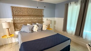 Un dormitorio con cama, ventilador de techo, mesita de noche con lámpara y ventana con cortinas.