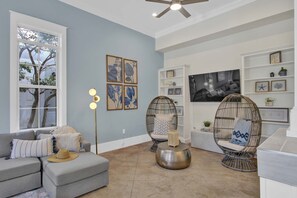 Relax in this inviting living room with a sofa, wicker chairs, and a coffee table, surrounded by framed artworks and a large window with a view of trees.