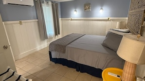 A hotel room with a bed, a small yellow table with a lamp, an air conditioning unit, and a window with curtains.