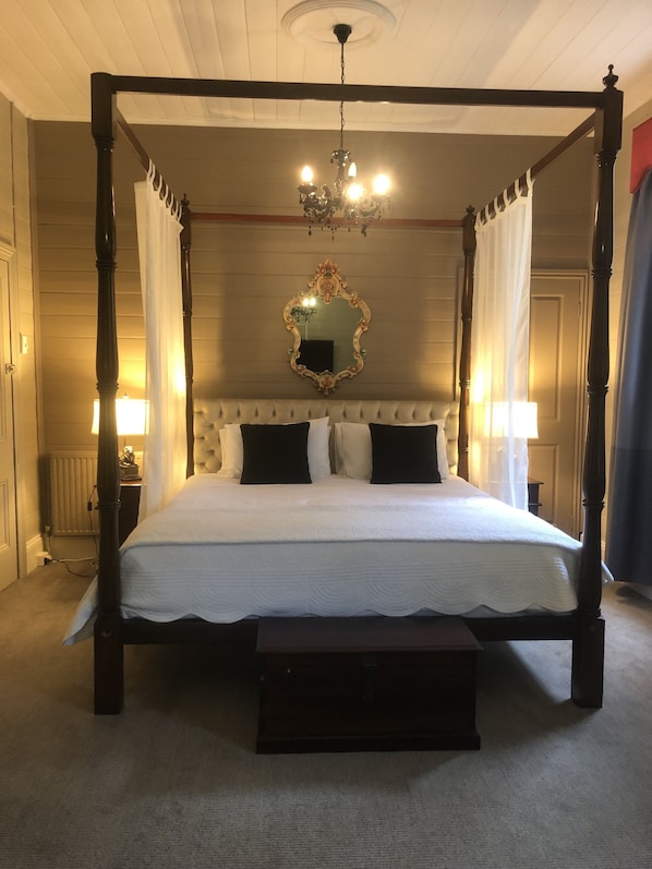 Classic four-poster bed with a canopy, adorned with a white bedspread and black pillows, sits beneath a large ornate mirror and a chandelier.