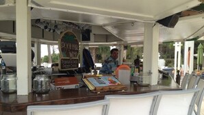 Un espacio de estar al aire libre cubierto con una mesa de madera y sillas blancas, un cartel publicitando bebidas tropicales, y una persona al fondo.