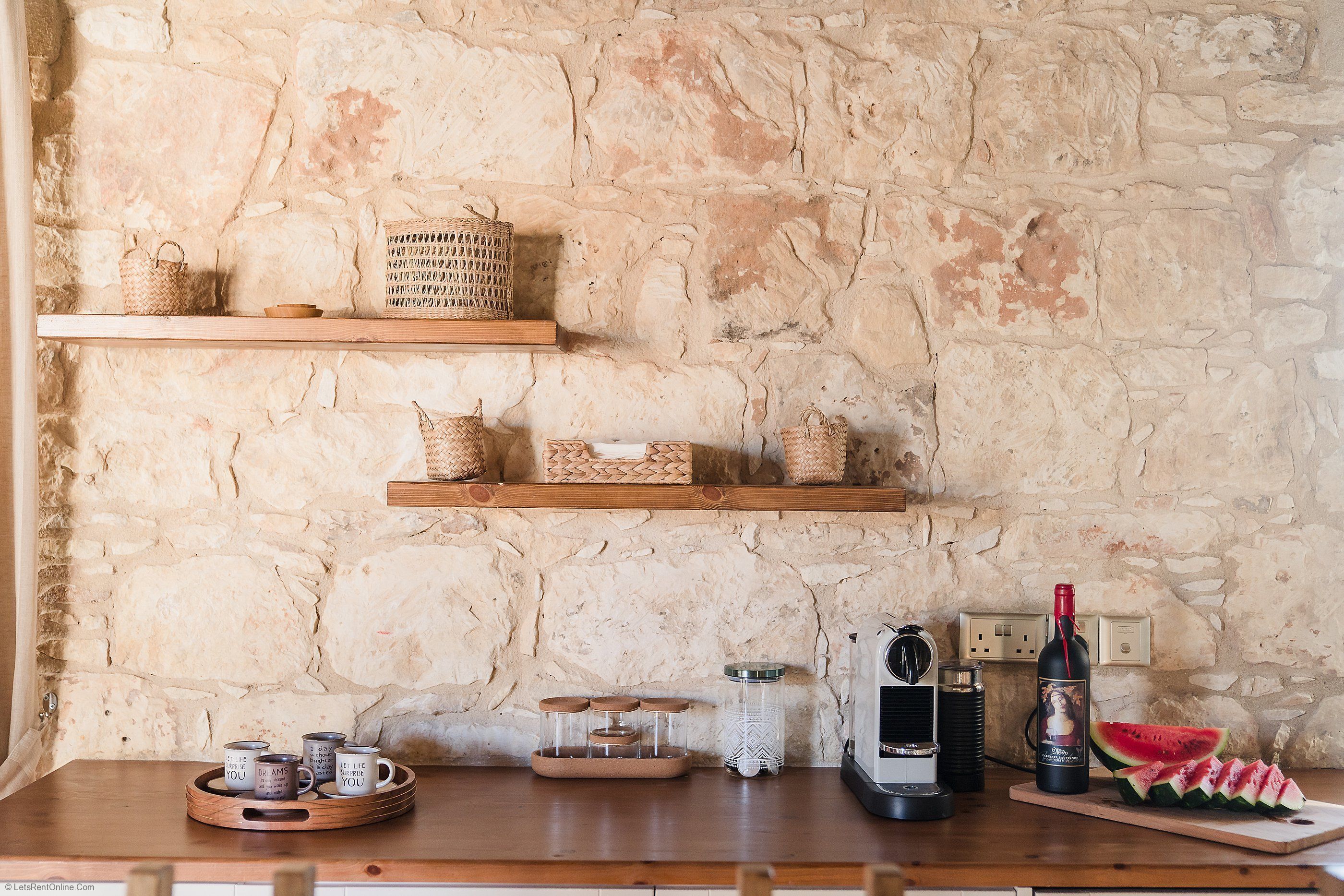 A rustic kitchen with stone walls, wooden shelves, and various kitchen items including a wine bottle, a watermelon, and a coffee setup.