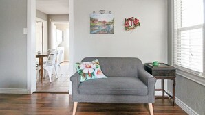 Un salon avec un canapé gris, une table à manger avec des chaises, une petite table d’appoint et une fenêtre avec des stores.