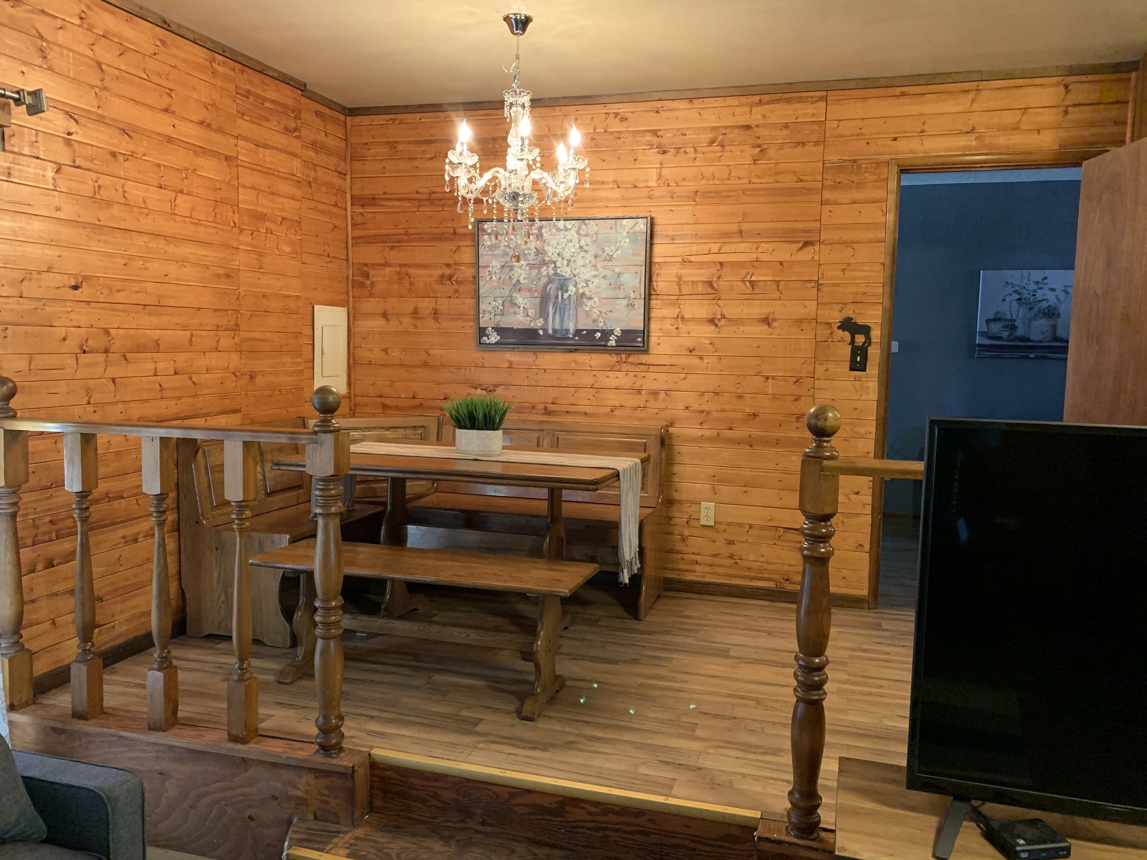 Un interior de madera con una mesa de comedor, bancos, una araña y un televisor.