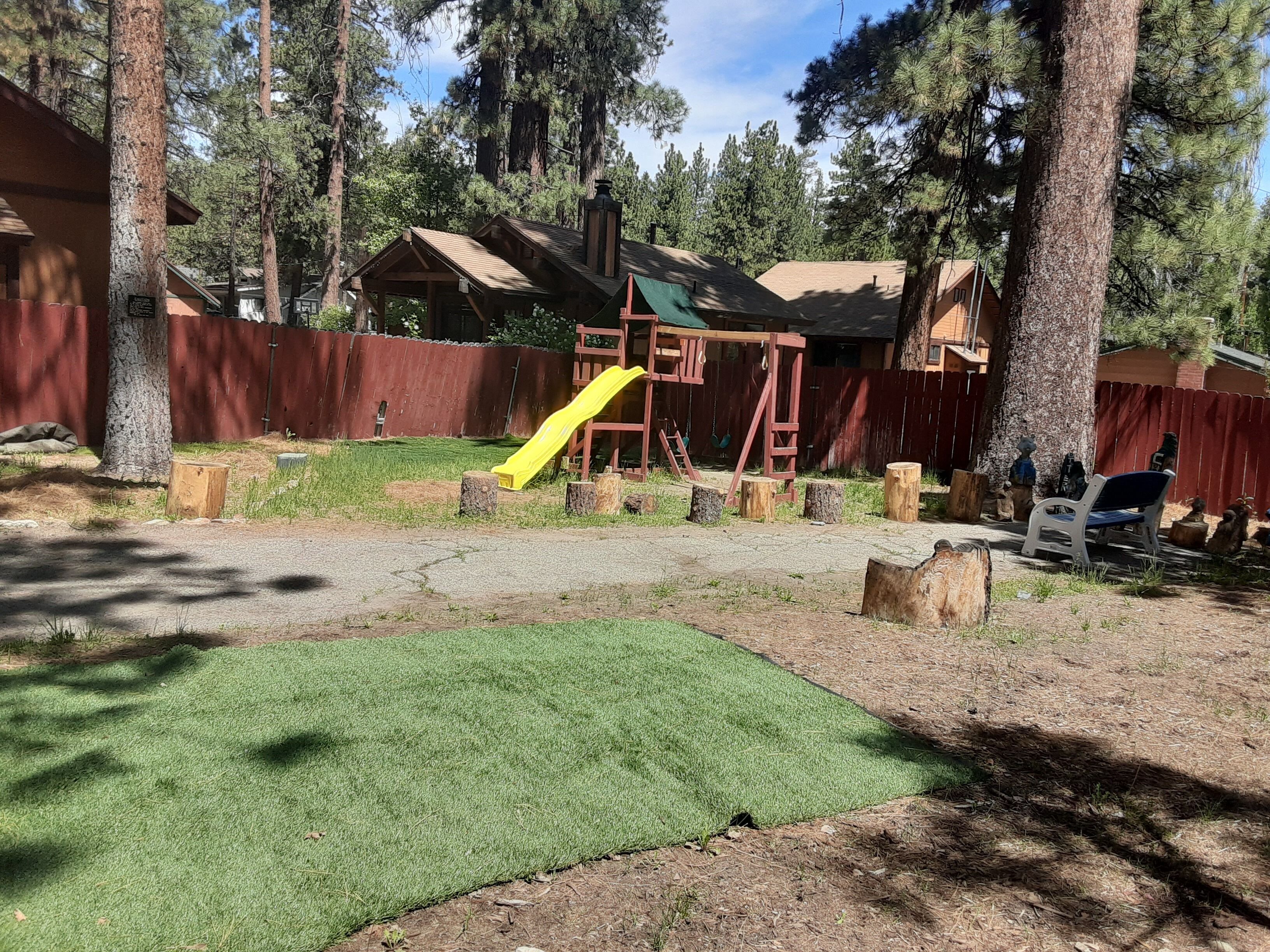 A playground with a yellow slide, wooden play structures, and a fenced area with trees.