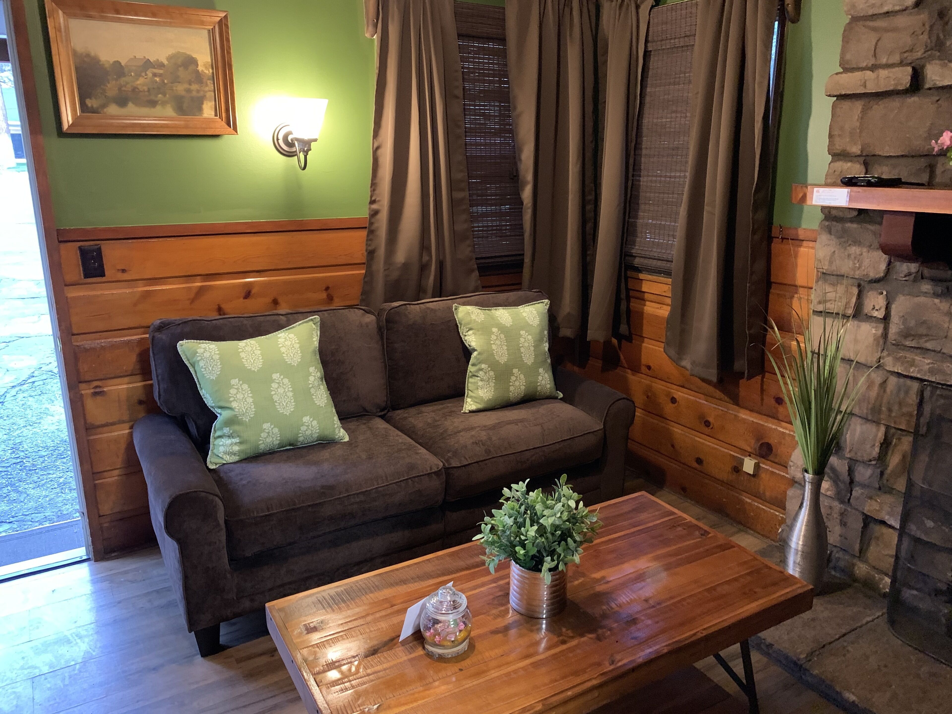 Una habitación acogedora con un sofá de madera, dos cojines estampados, una mesa de centro de madera con una planta, una chimenea de piedra y un cuadro en la pared.