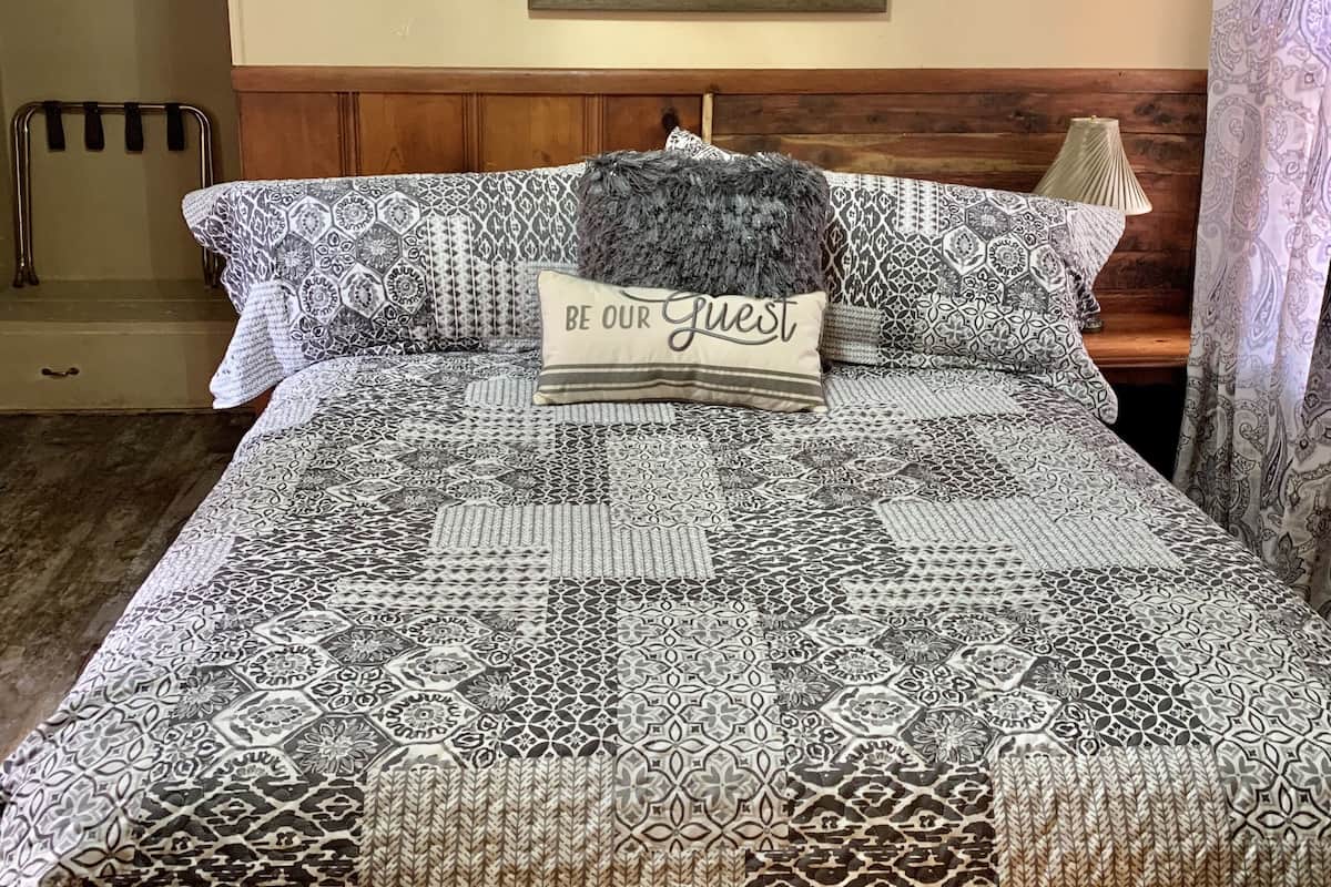 A neatly made bed with a patterned bedspread, a pillow with 'Be Our Guest' text, and a mirror above the bed.
