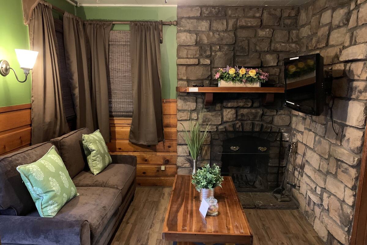 A cozy room with a stone fireplace, a wooden sofa with green cushions, a wooden coffee table, and a TV mounted above the fireplace.