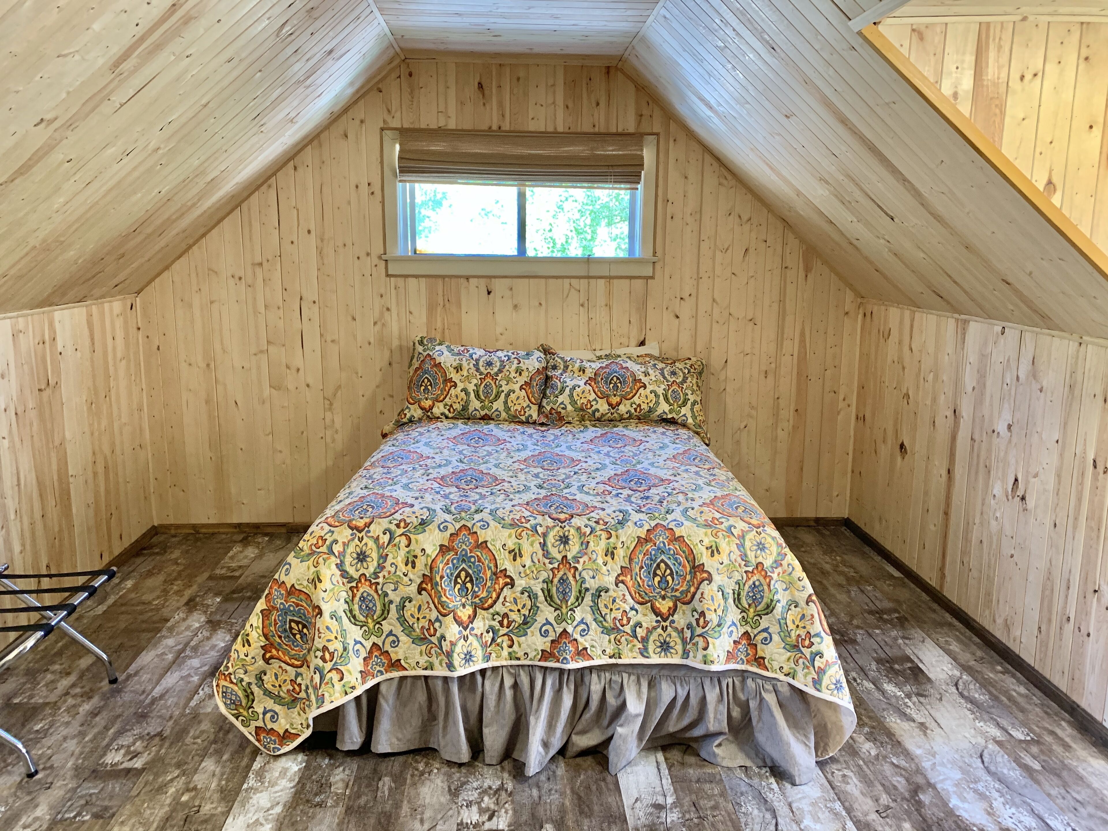 Una camera da letto mansardata accogliente, con travi a soffitto in legno, un letto grande con copripiumino fantasia e una piccola finestra vista sul verde.