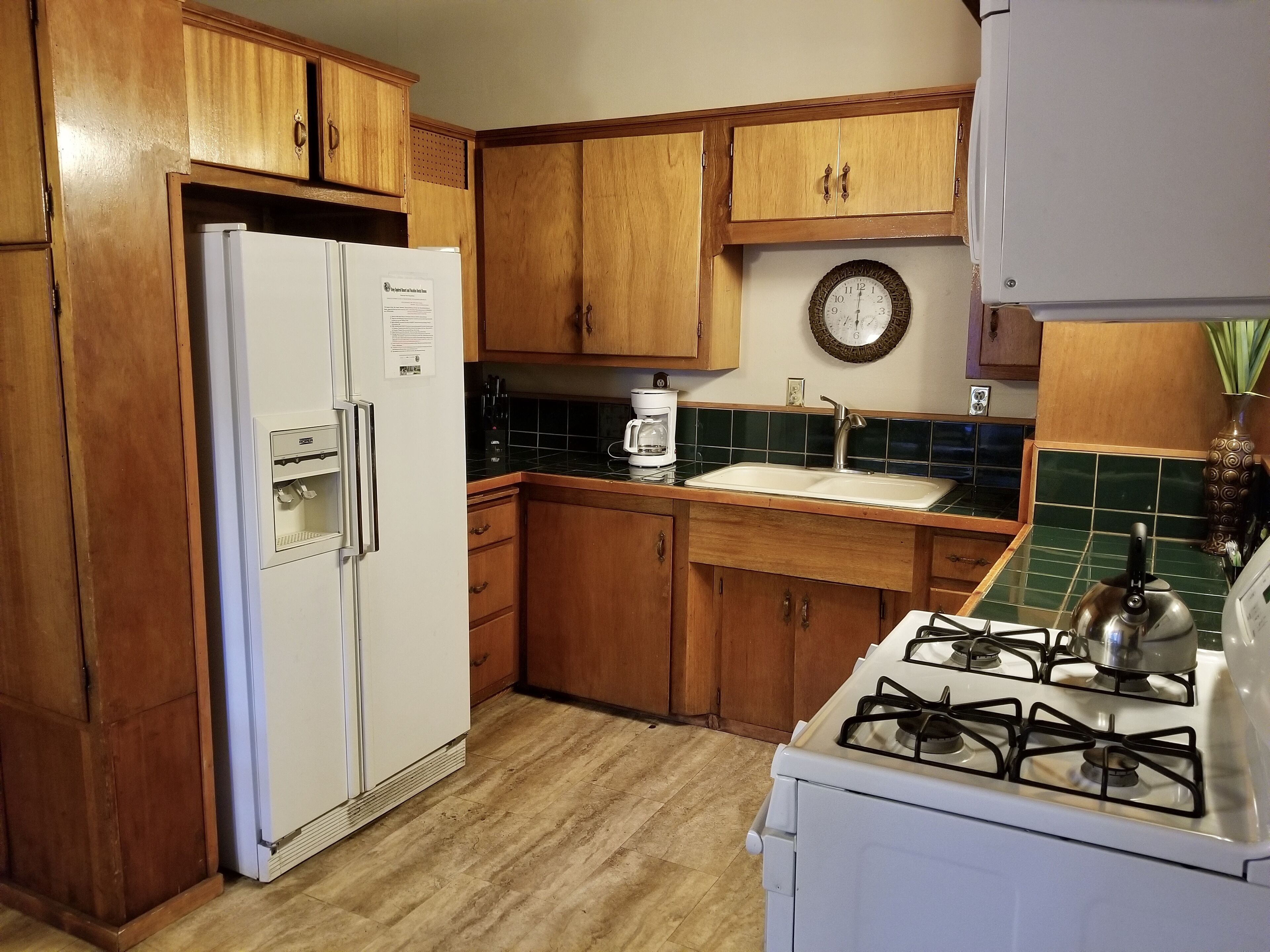 A kitchen with wooden cabinets, a white refrigerator, a stove, a sink, and a clock on the wall.