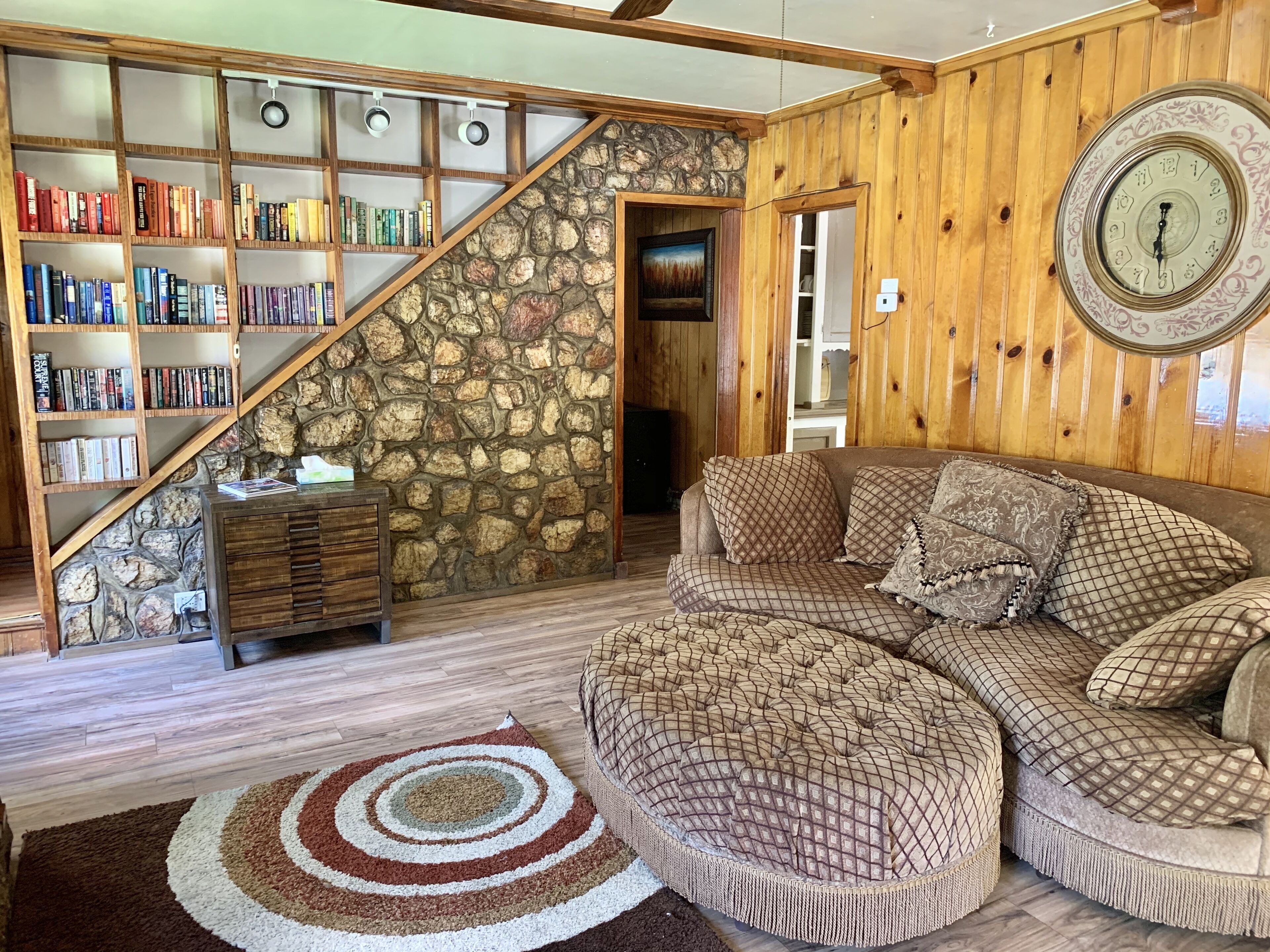 A cozy living room with a stone wall, wooden bookshelves, a large clock, and a sectional sofa.