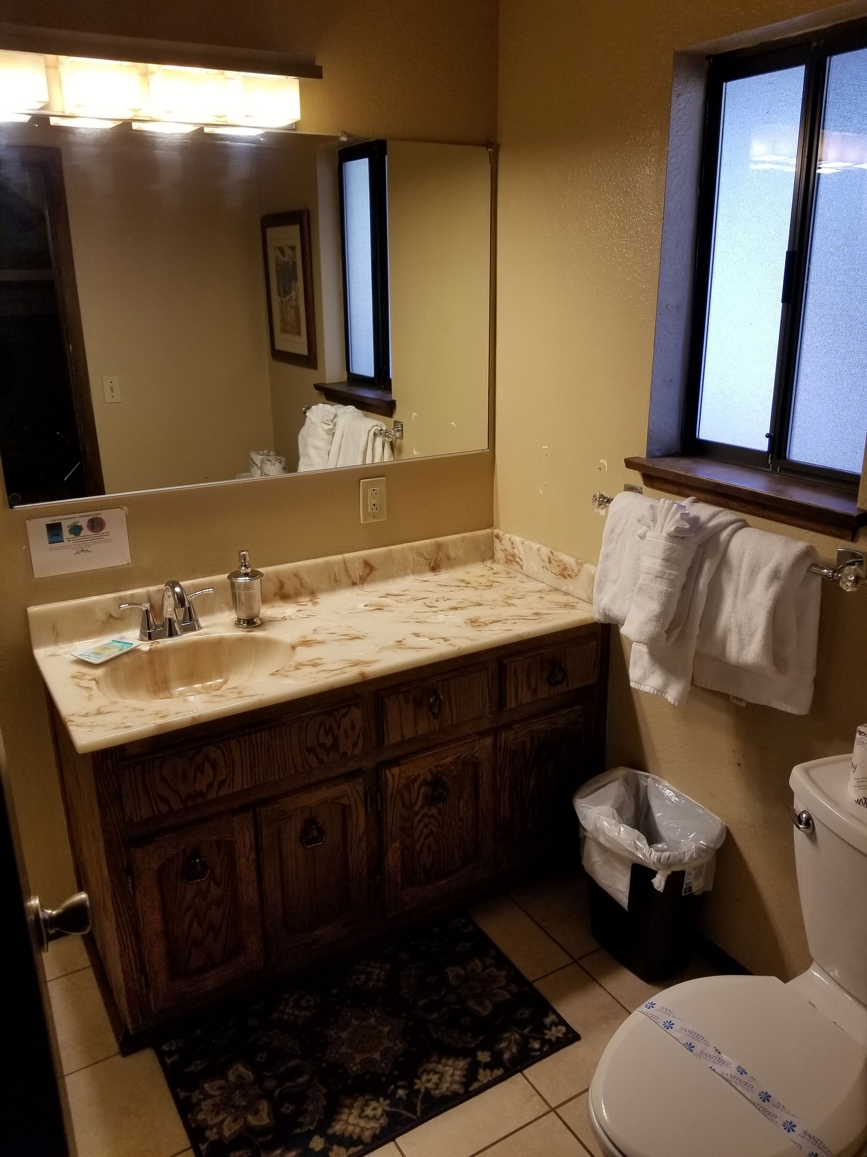 A bathroom with a marble sink, wooden vanity, a mirror, a window, a toilet, and a trash bin.