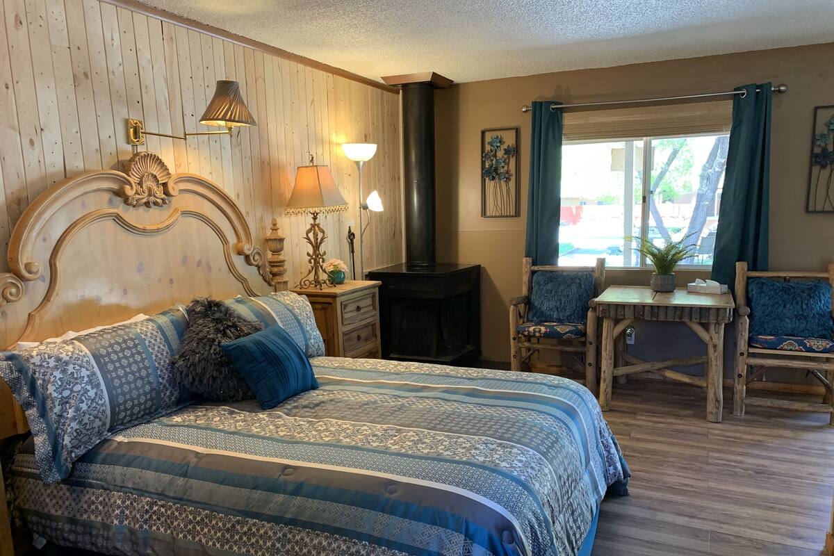 A bedroom with a large bed, two bedside tables, a wooden headboard, a small table with a plant, and a window with curtains.