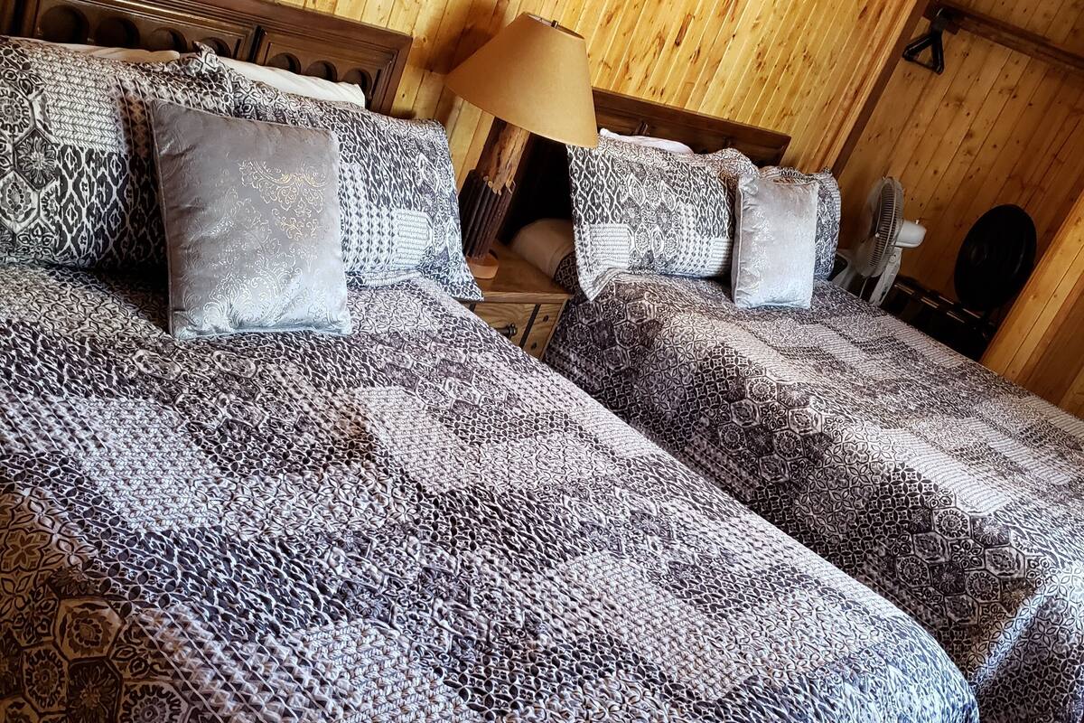 A room with two beds, each with patterned bedding and a wooden headboard. A lamp and a fan are visible on the nightstand.