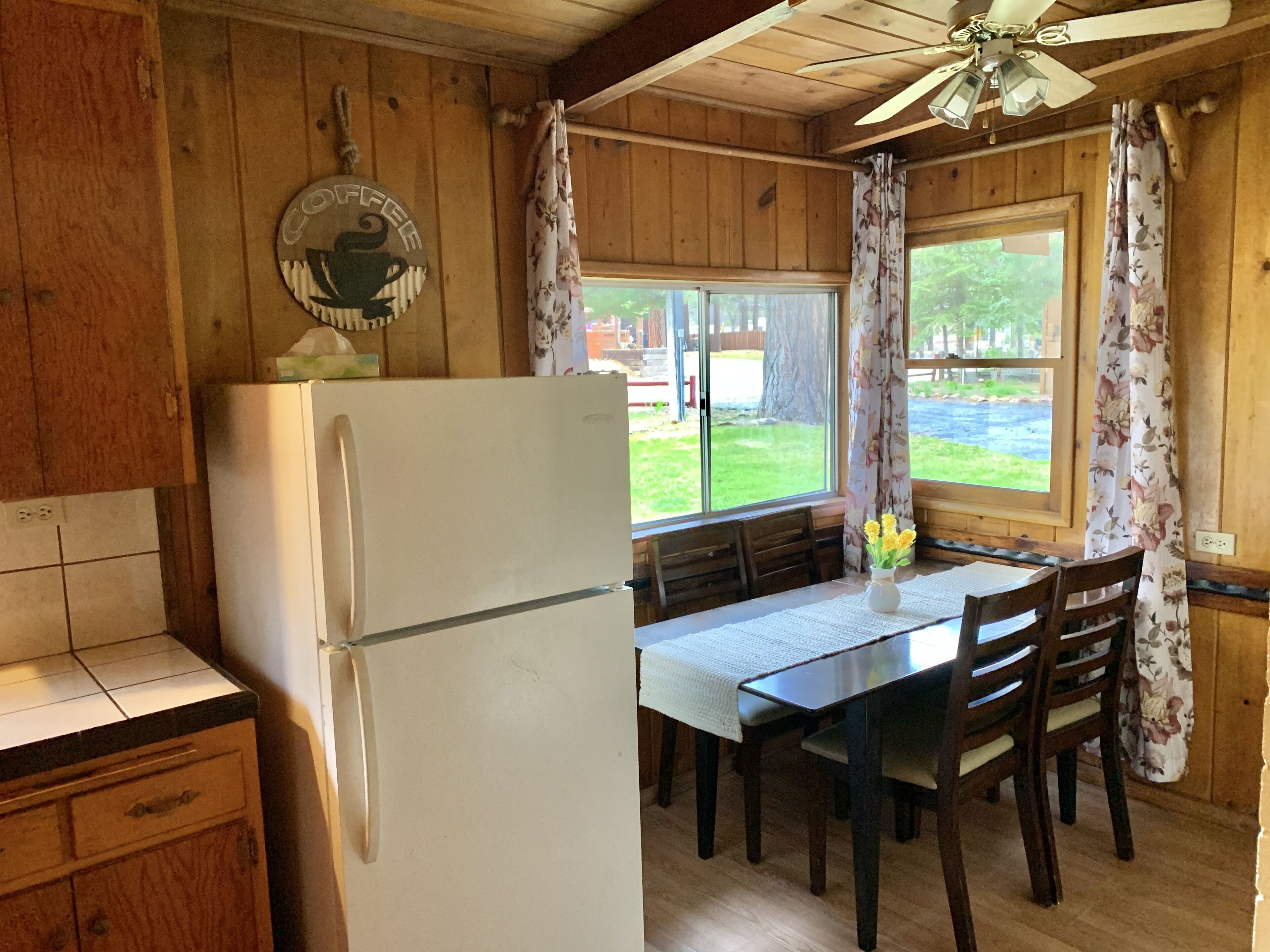 A kitchen with wooden walls, a white refrigerator, a dining table with chairs, and a window with curtains.