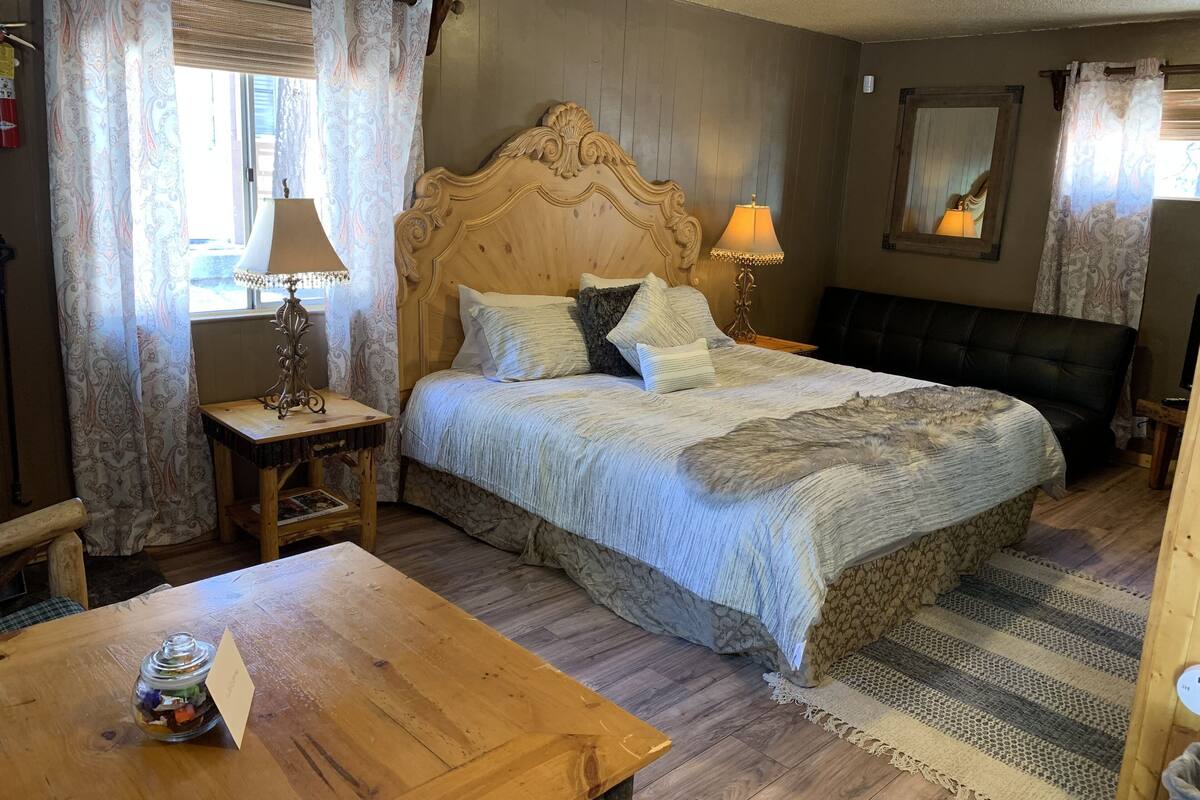 A cozy bedroom with a wooden bed, two bedside lamps, a wooden nightstand, a small table with a jar of snacks, and a stone wall.