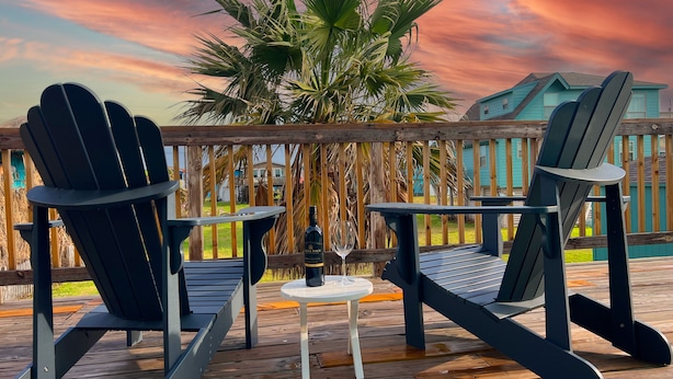 Une terrasse en bois avec deux fauteuils Adirondack et une petite table, une bouteille de vin et un verre posés sur la table.