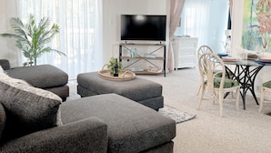 Un salon avec des canapés gris, un téléviseur, une table à manger avec des chaises, un ventilateur de plafond et une grande fenêtre ornée de rideaux légers.