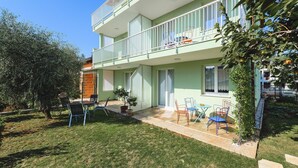 A two-story building with a balcony, a small patio area with chairs and a table, and a garden with a tree and grass.