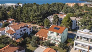 A residential area with houses featuring red-tiled roofs, surrounded by dense greenery and a view of the sea.