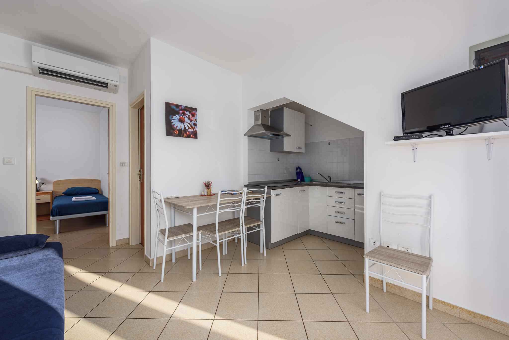 Ein kompakter Raum mit Bett, Esstisch mit Stühlen, einer Küchenzeile und einem an der Wand befestigten Fernseher.
