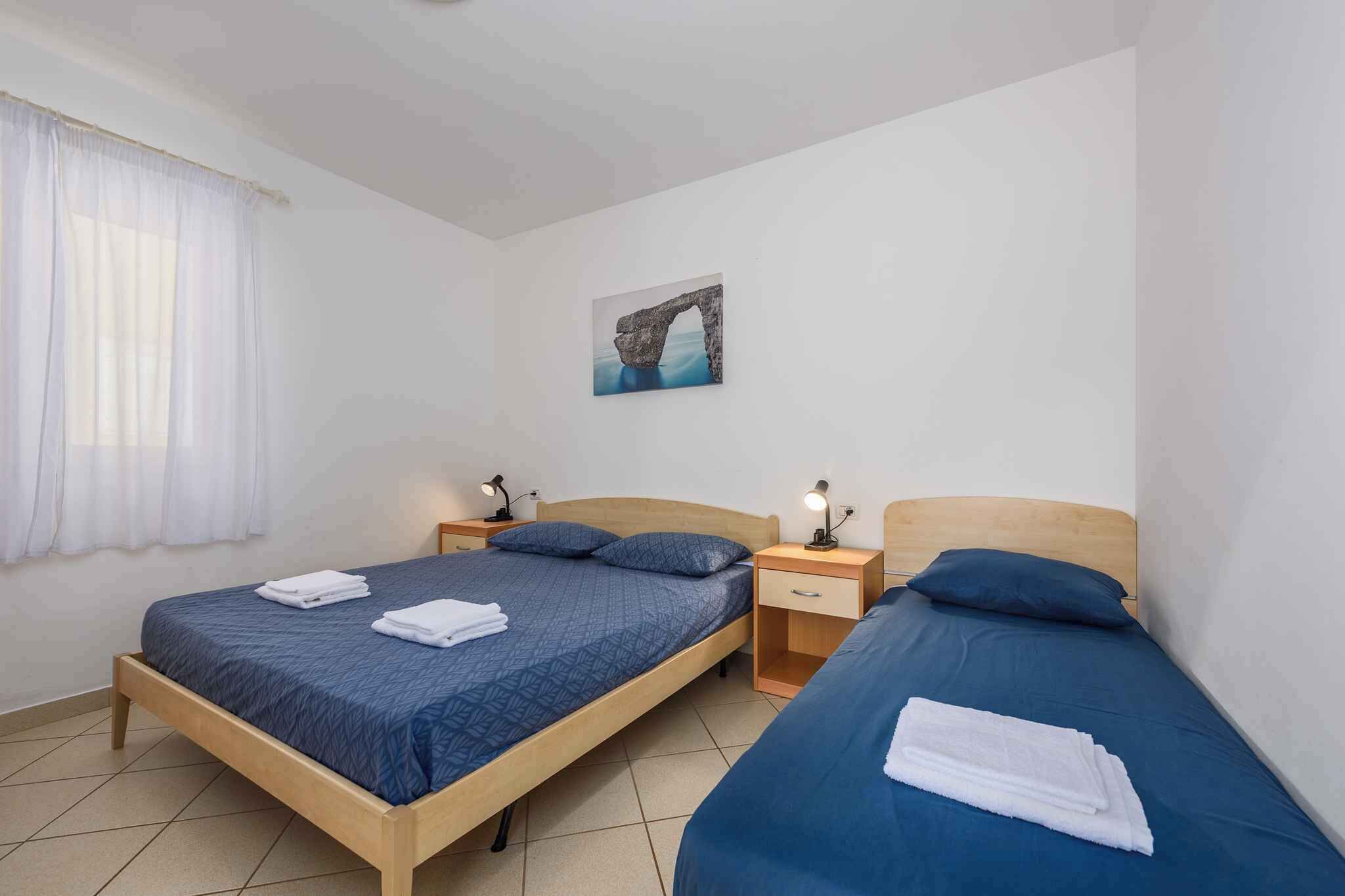 A room with two beds, each with a blue bedspread and white towels. There is a painting of a stone arch on the wall, and a window with white curtains.