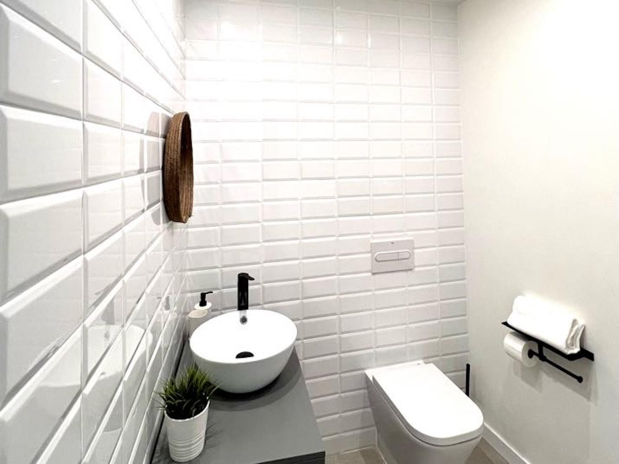 Un baño moderno con azulejos blancos tipo metro, un lavabo redondo, un grifo negro, una pequeña planta en maceta, un inodoro y un perchero para toallas.