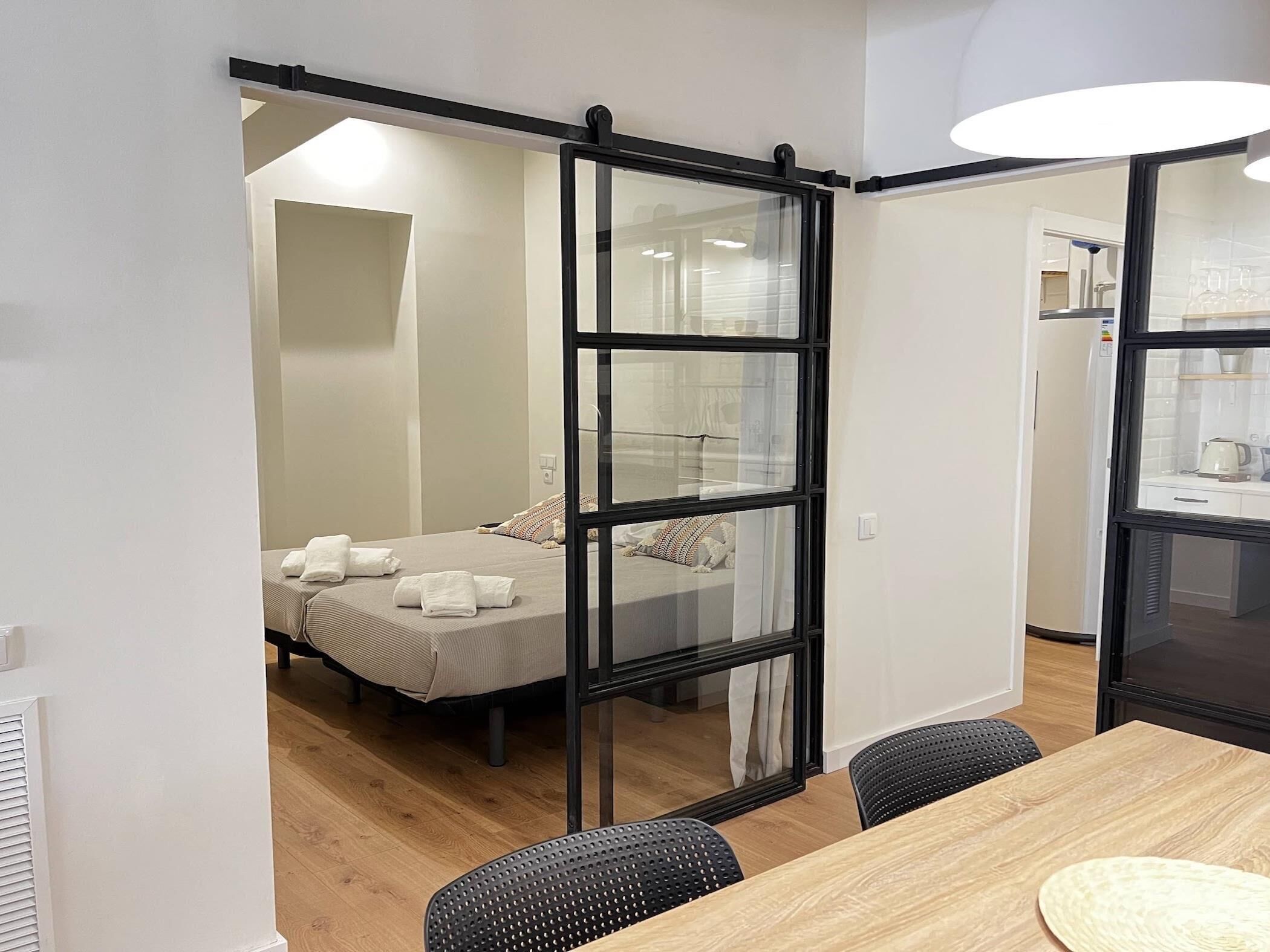 Een moderne hotelkamer met een schuifdeur naar een badkamer, een houten eettafel met stoelen en een bed met netjes opgevouwen handdoeken.