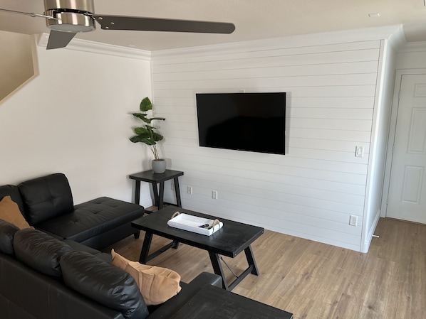 A modern living room with a black leather sofa, a small black table, a potted plant, and a mounted TV on a white paneled wall.