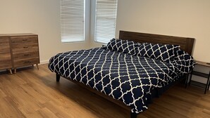 A bedroom with a bed, wooden dresser, and two windows with blinds.