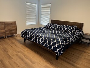 A bedroom with a bed, wooden dresser, and two windows with blinds.