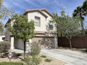 A single-story house with a beige exterior, a front garage, and a small front yard with trees and shrubs.