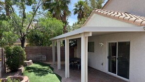 A covered patio area with a brick border, a small lawn, and a building with a tiled roof and sliding glass doors.