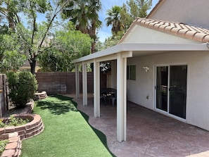A covered patio area with a brick border, a small lawn, and a building with a tiled roof and sliding glass doors.