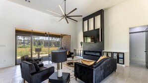 Ein modernes Wohnzimmer mit Kamin, schwarzen Sofas und einem großen Fenster mit Blick auf einen Wald.