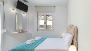 Ett välordnat sovrum med en säng, en sänglampa, en tv som är monterad på väggen och ett fönster med utsikt över omgivningen.
