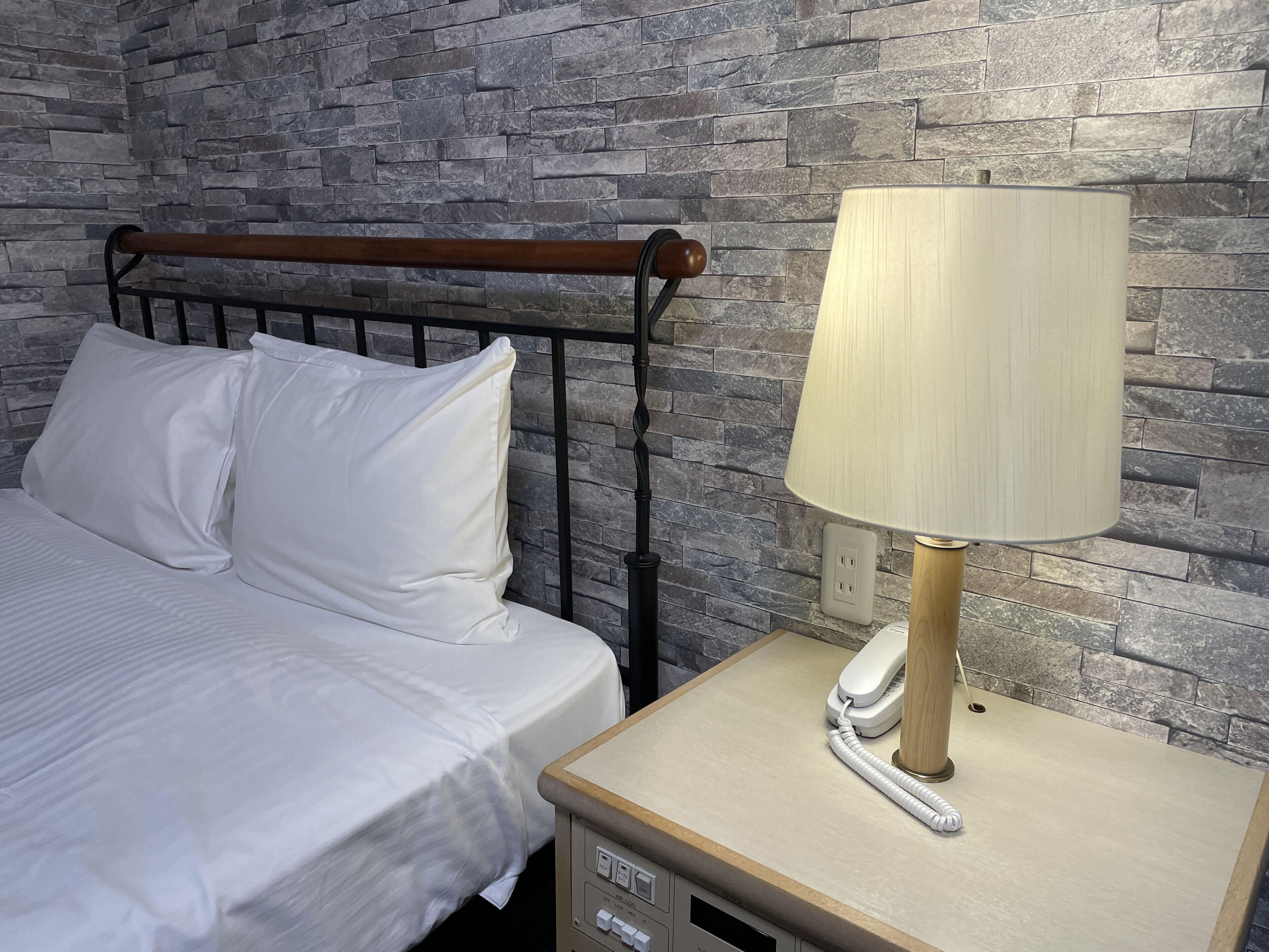 Una cama con ropa de cama y almohadas blancas, un cabecero de madera y una mesita de noche con una lámpara y un teléfono.