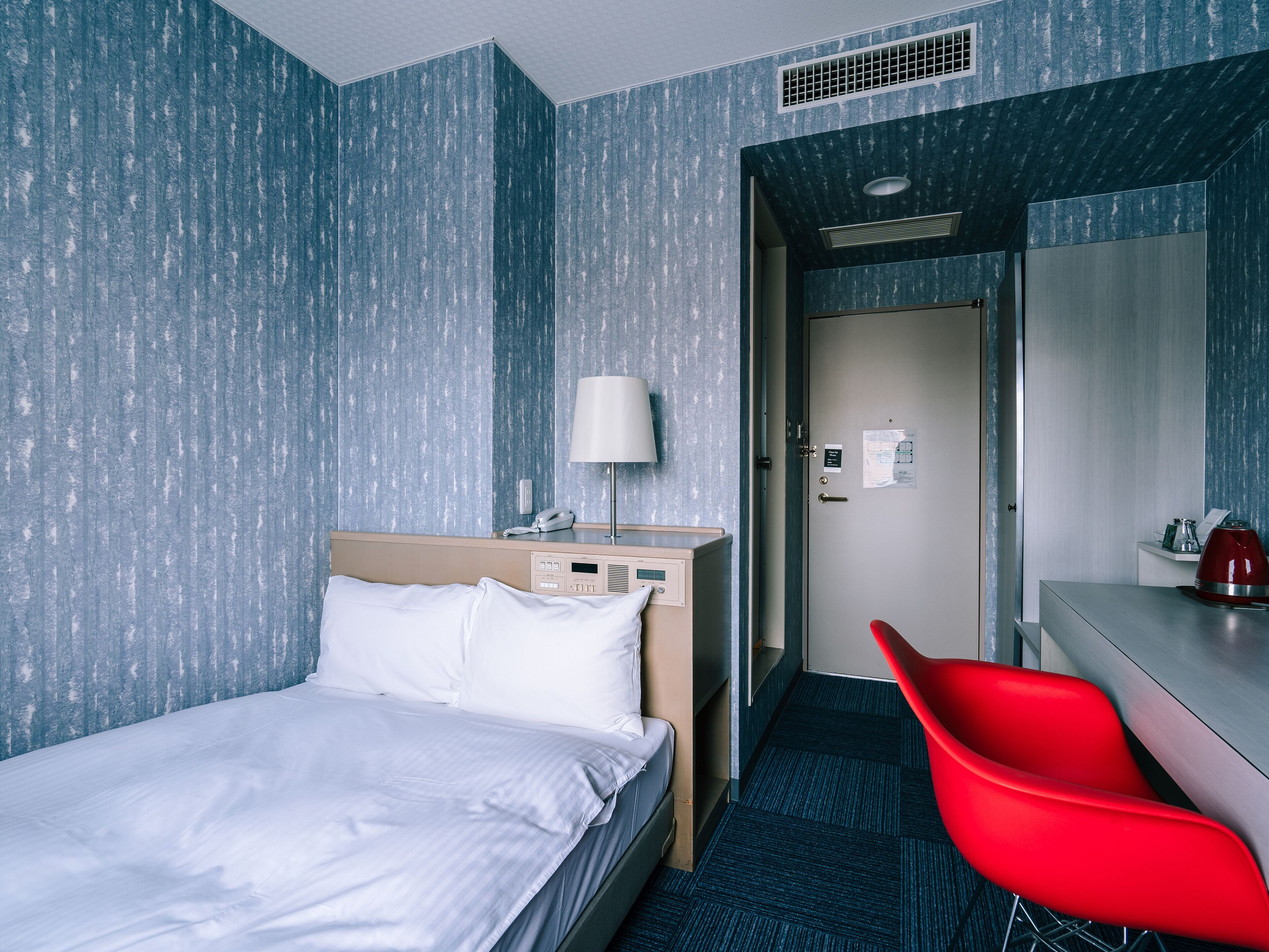 A hotel room with a bed, a desk with a red chair, a lamp, and a bathroom visible through an open door.
