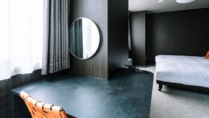 A modern hotel room with a dark wood headboard, a round mirror, a bed with white bedding, and a dark wooden desk with a chair.