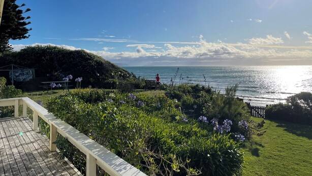 A coastal view with a wooden deck, lush greenery, and a person standing by the water. - Absolute beachfront Raumati (Paraparaumu)