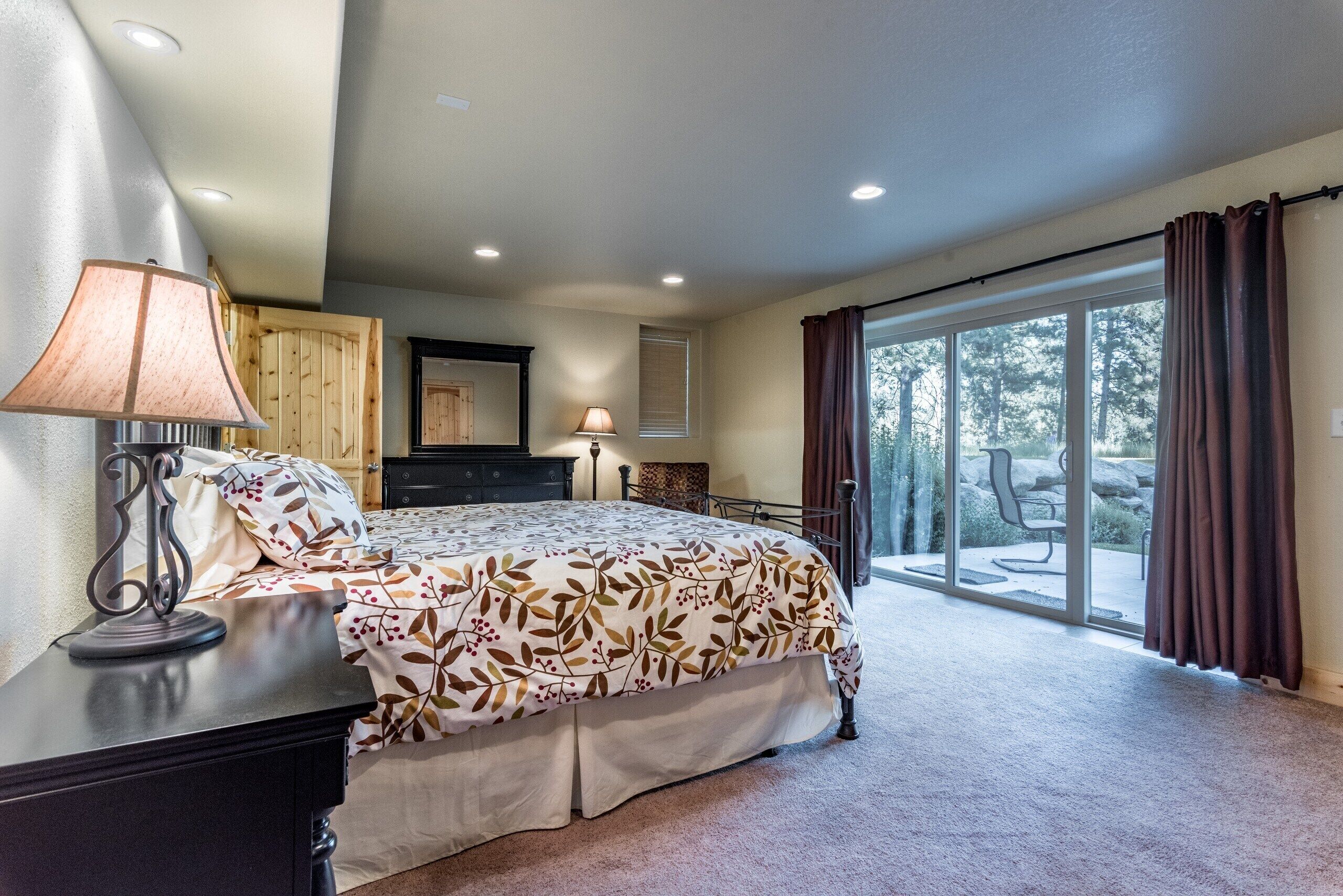 A bedroom with a large bed, a desk with a lamp, a dresser, and a sliding glass door leading to an outdoor area.