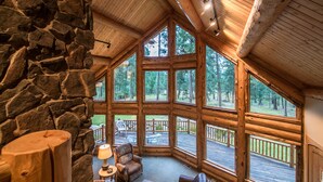 A log cabin interior with a stone wall, large windows, a ceiling fan, and a cozy seating area. - Pineview Lodge (Hamilton)