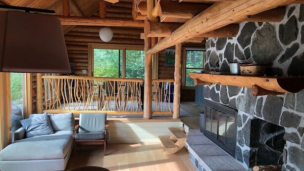 Interior acolhedor de uma cabana, com lareira de pedra, vigas de madeira no teto e um sofá perto da janela.