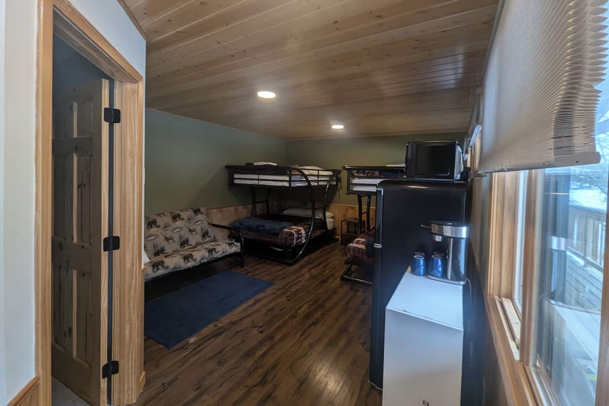 Een compacte kamer met stapelbedden, een slaapbank, een kleine kitchenette en een raam met zonwering.