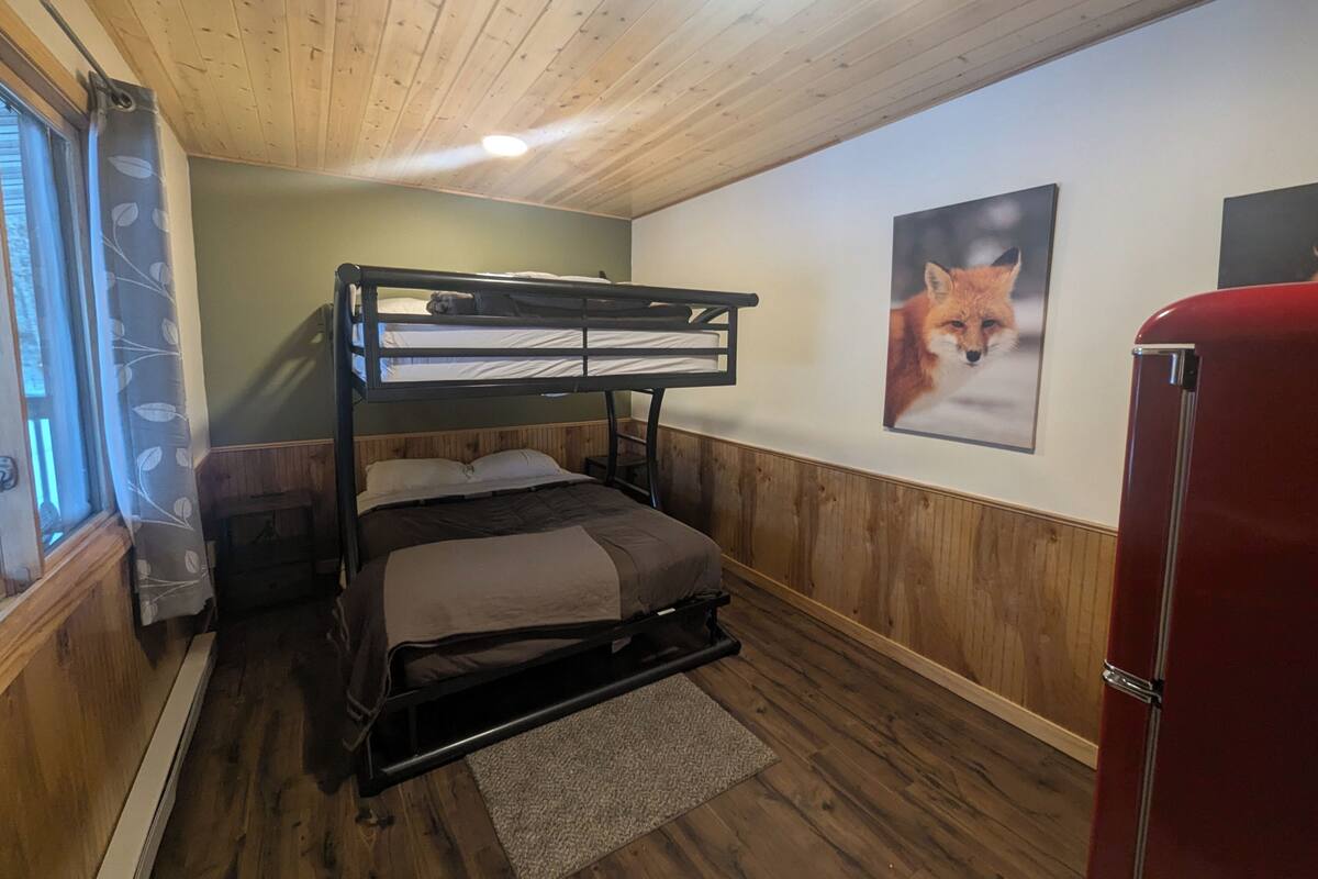 Een slaapkamer met stapelbed, houten plafond, raam met gordijn met bladmotief, een vosposter aan de muur en een rode kast.