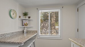 Een keuken met granieten werkbladen, witte keukenkastjes, een klok en een raam met jaloezieën.