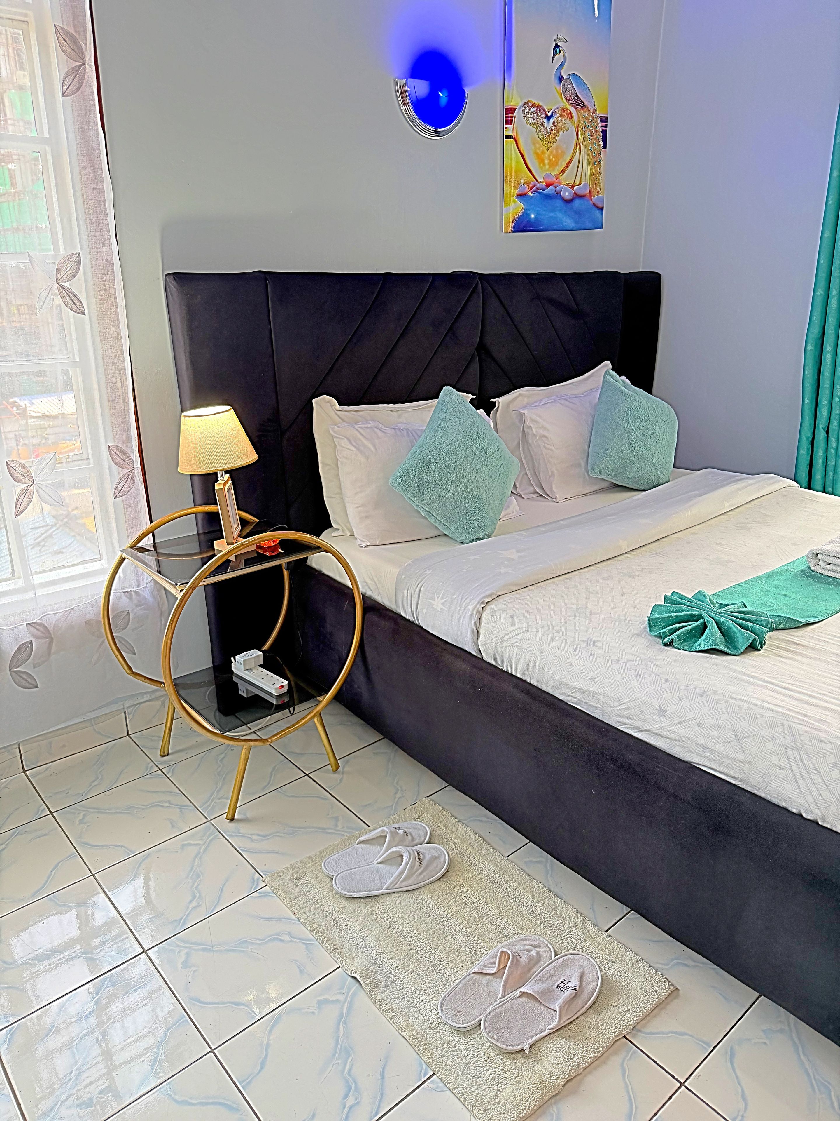 Une chambre à coucher avec un lit, une table de chevet avec une lampe, un élément de décoration murale et une fenêtre avec des rideaux.